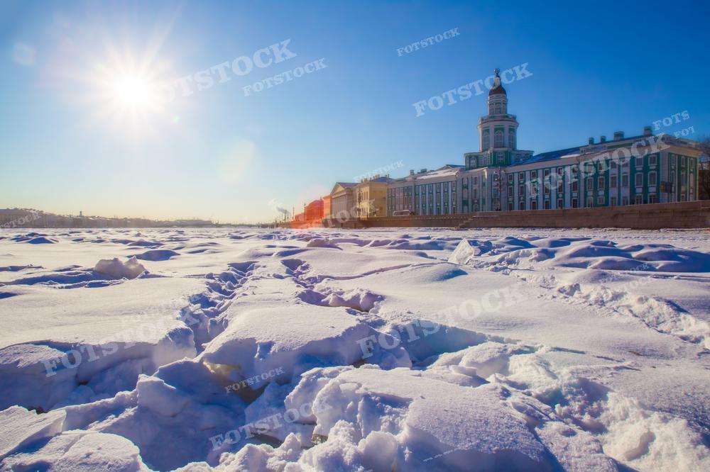 На фотографии изображён зимний вид Санкт-Петербурга с застывшей поверхностью реки Невы, покрытой снегом. На заднем плане виден классический архитектурный ансамбль города, включающий историческое здание Кунсткамеры с колоннадой и куполом. Солнце стоит низко над горизонтом, создавая яркую бликовую засветку и длинные тени на снегу. Это фото может использоваться для иллюстрации туристических и культурных статей о Санкт-Петербурге, в качестве зимнего пейзажа для календарей или открыток, а также для образовательных материалов о России.
